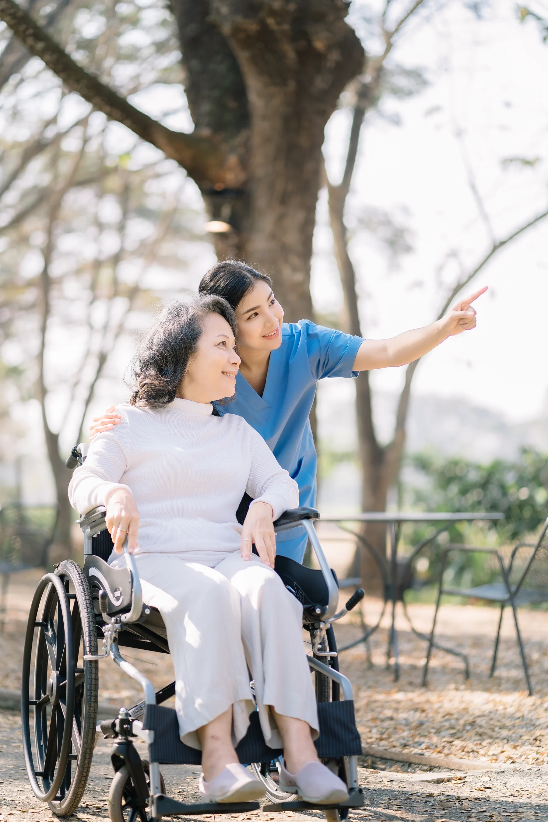 Caregiver in blue scrubs pointing while standing beside an older woman in a wheelchair outdoors